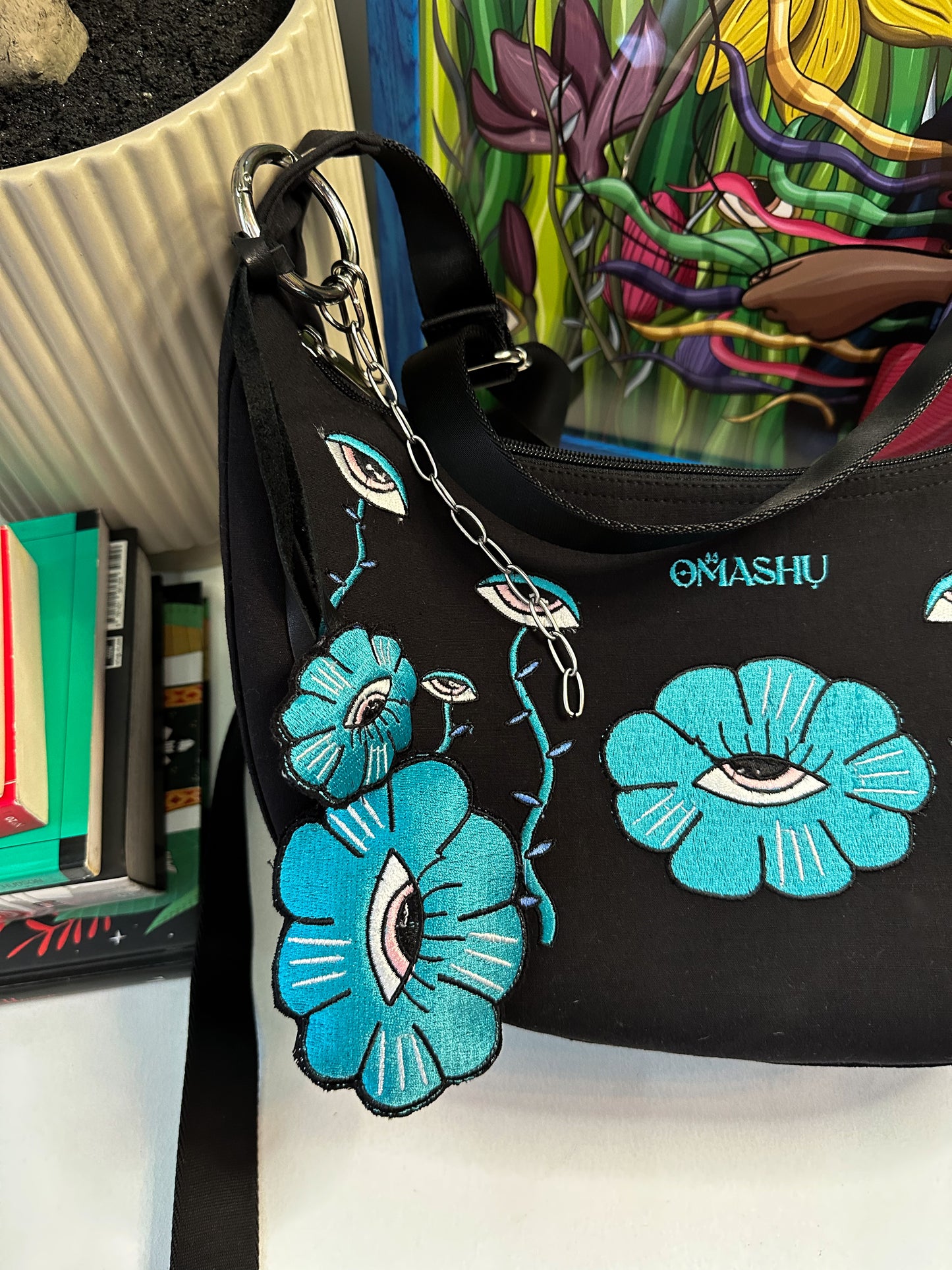 a bag and key chain both has embroidered illustration, it also has OMASHU branding on it. on the back ground there is a pot and books