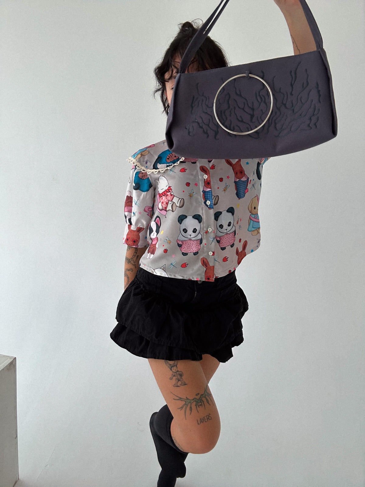 A person is wearing a colorful blouse with cartoon animal designs, including pandas and rabbits, along with a black ruffled skirt. The person is holding up a dark grey handbag with a large metal ring accent, obscuring part of their face. The playful outfit is paired with knee-high black socks, creating a fun and stylish combination. Visible tattoos on the legs add a unique detail to the overall look.