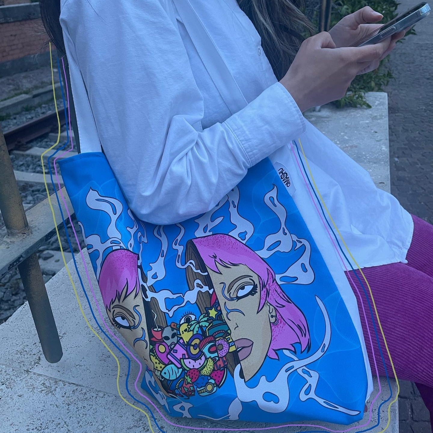 A person wearing a white shirt and pink pants, sitting on a bench and holding a phone in their hand. The person is wearing a blue tote bag with a colorful design of two faces, one pink and one blue, with a bunch of colorful objects in between them. The background consists of a brick wall and a metal railing. tag says 'OMASHU'.