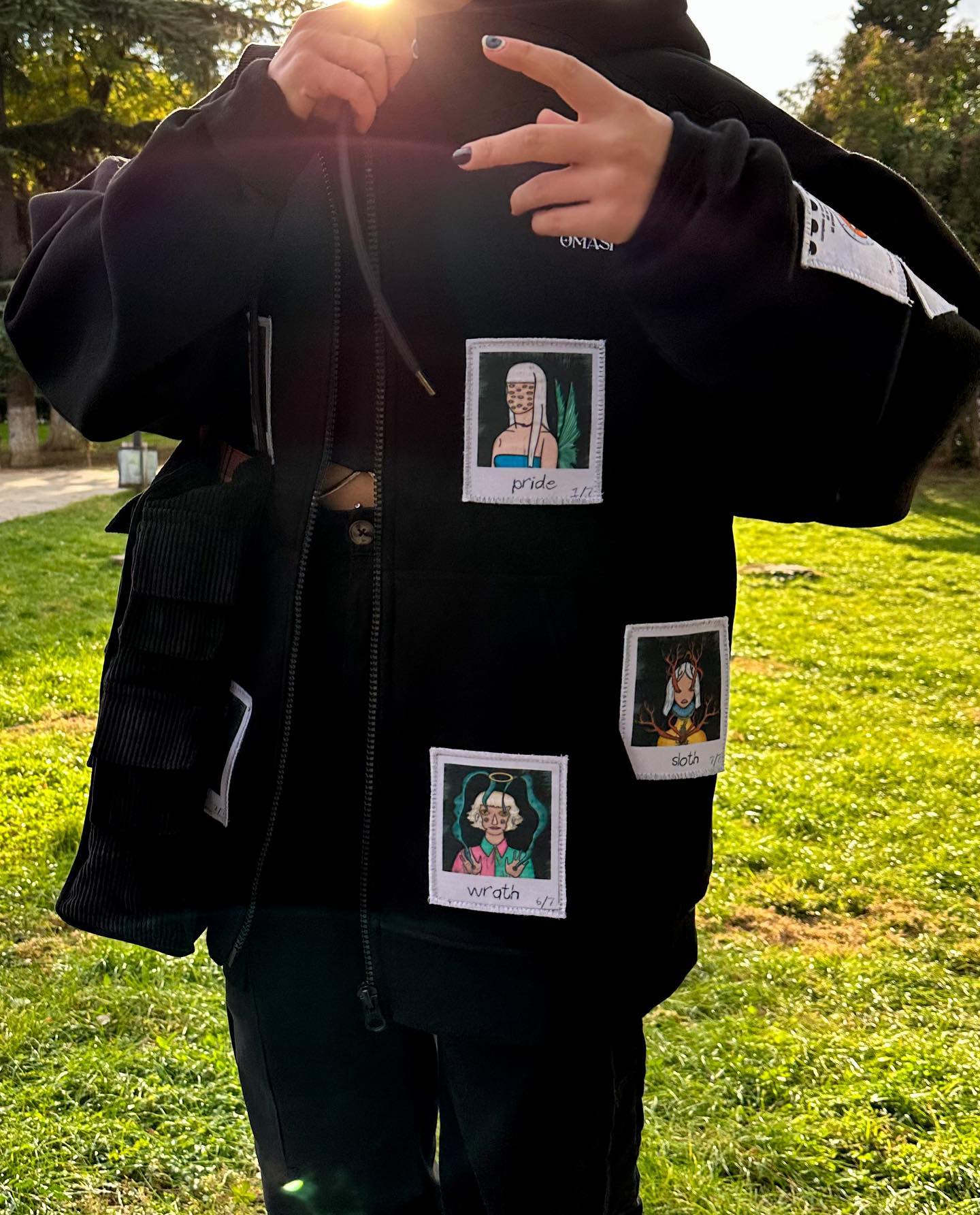 Black hoodie with three square patches on it. The patches are white with a black border and have illustrations of people’s faces and text on them. The top patch has a person with long hair and the text ‘pride’. The middle patch has a person with short hair and the text ‘wrath’. The bottom patch has a person with curly hair and the text ‘sloth’