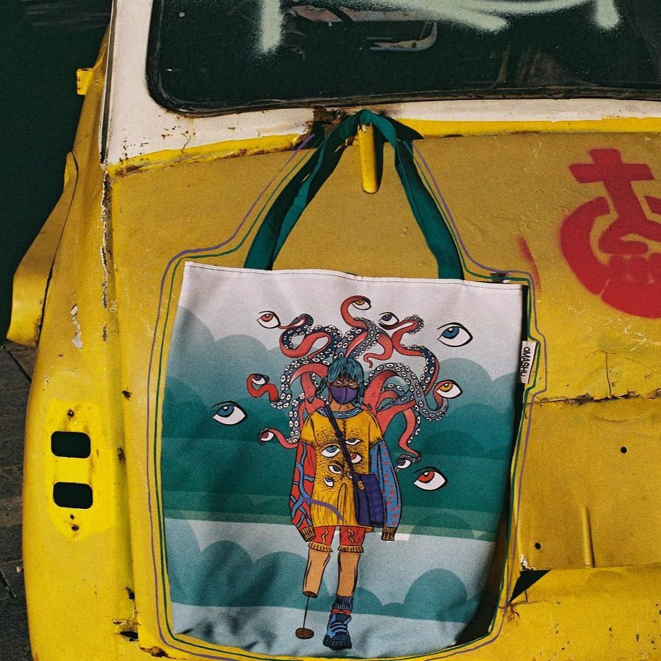 A tote bag hanging on the side mirror of a yellow car. The tote bag has a colorful illustration of a person with an octopus on their head. The person is wearing a blue and orange outfit with a red scarf. The octopus has red tentacles that are reaching out in all directions. The background of the tote bag is a blue-green color with white clouds. The car is an old model with rust and peeling paint. The car has a red character on the side.”