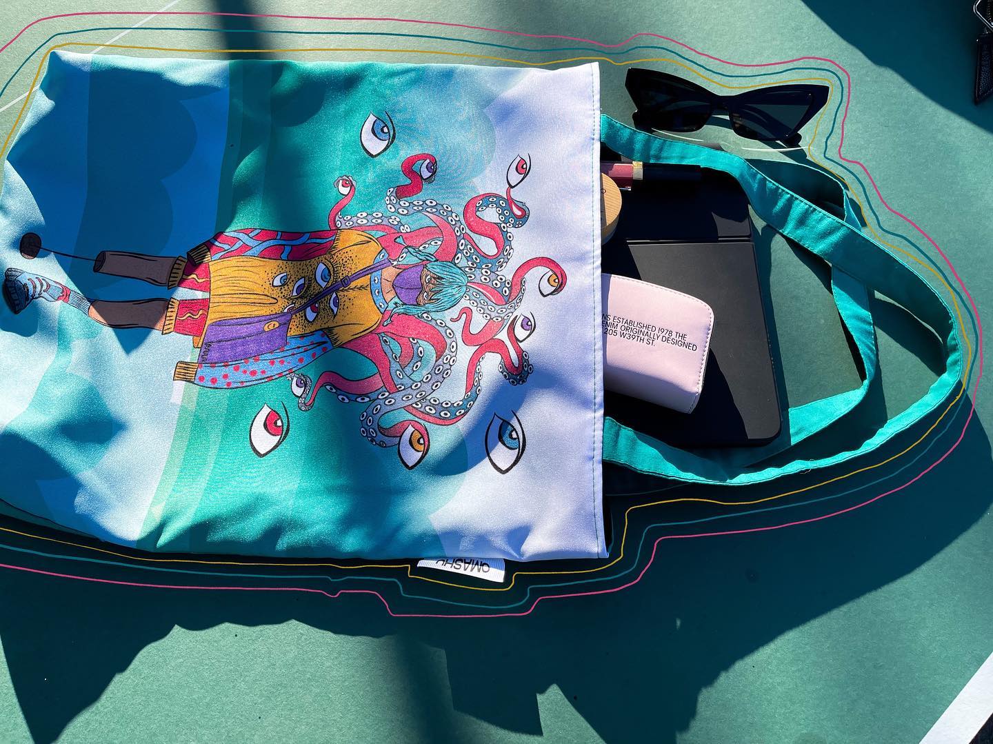 A blue tote bag with a colorful octopus design on it. The octopus is pink and purple with blue eyes and tentacles. The bag is lying on a green table with a pair of sunglasses and a black phone next to it. The phone has a pink case with ‘I love you’ written on it. The table is in an outdoor setting with sunlight shining on it.