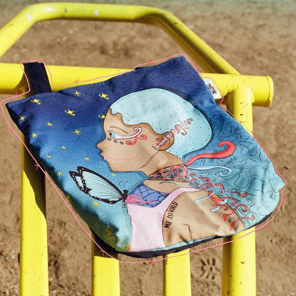 A photo of a colorful OMASHU tote bag with a blue strap, featuring a design with a woman’s face in profile wearing a blue headscarf, a butterfly, and a red ribbon, hanging on a bright yellow metal railing that is slightly rusted, against a sandy ground background with small rocks scattered about.