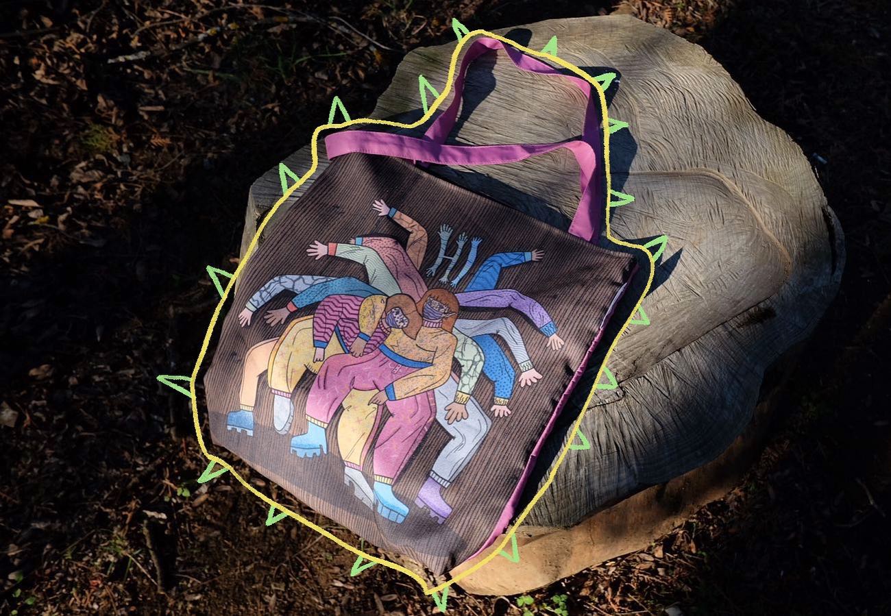 A photo of a colorful tote bag made of brown fabric with a purple handle and green and yellow zigzag borders, featuring a design of a group of people with their arms stretched out in a circle, wearing pink, orange, and yellow clothes, on a tree stump in a forest with fallen leaves, taken during the day with natural lighting.