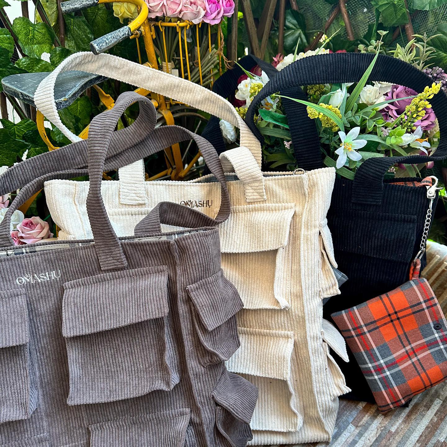 A collection of bags in different colors and styles arranged in a pile on a wooden surface with plants and flowers in the background. The bags are made of different materials such as corduroy, knit, and plaid. The bags have different handle styles such as round, square, and braided. The bags have different colors such as brown, beige, black, and orange. The bags have different sizes and shapes such as tote bags, shoulder bags, and crossbody bags. The bags have different brand label ‘OMASHU’