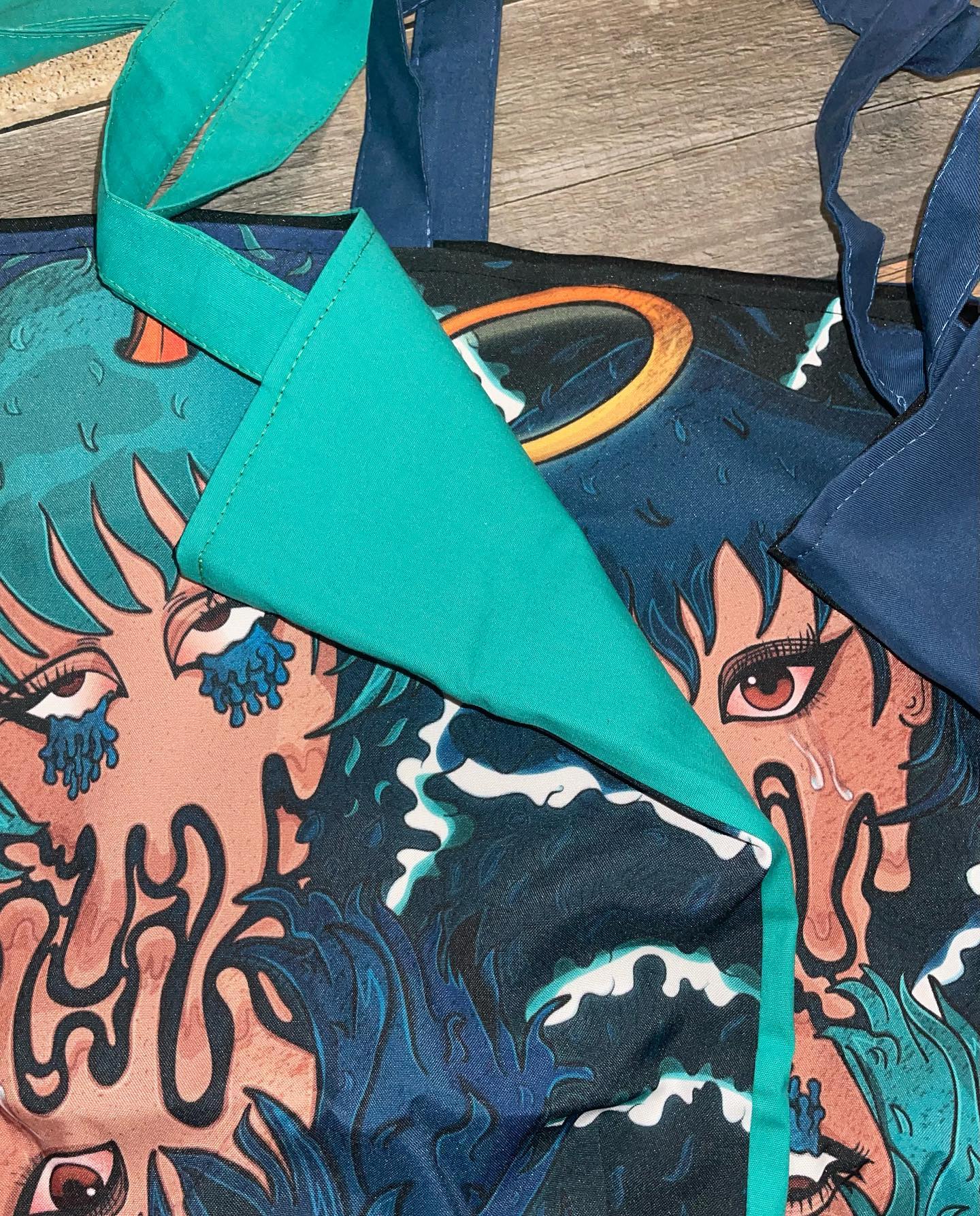 A blue and green tote bag with a graphic design on it. The graphic design is of a person’s face with a dragon-like creature on top of their head. The person’s face is in profile and they have a red eye. The dragon-like creature is blue and green with orange horns. The tote bag has blue straps and a green handle. The background is a wooden floor.