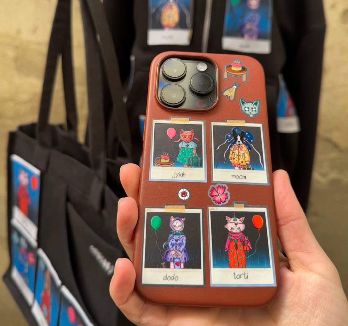 A close-up image of a brown smartphone case decorated with Polaroid-style patches featuring colorful cartoon animals from OMASHU. The case displays four characters: "Jiyah," "Mochi," "Dodo," and "Torti," each holding balloons or birthday-themed items. The background shows matching tote bags and hoodies with similar animal designs. The overall style is playful and whimsical, with bright colors and detailed illustrations of each animal character.