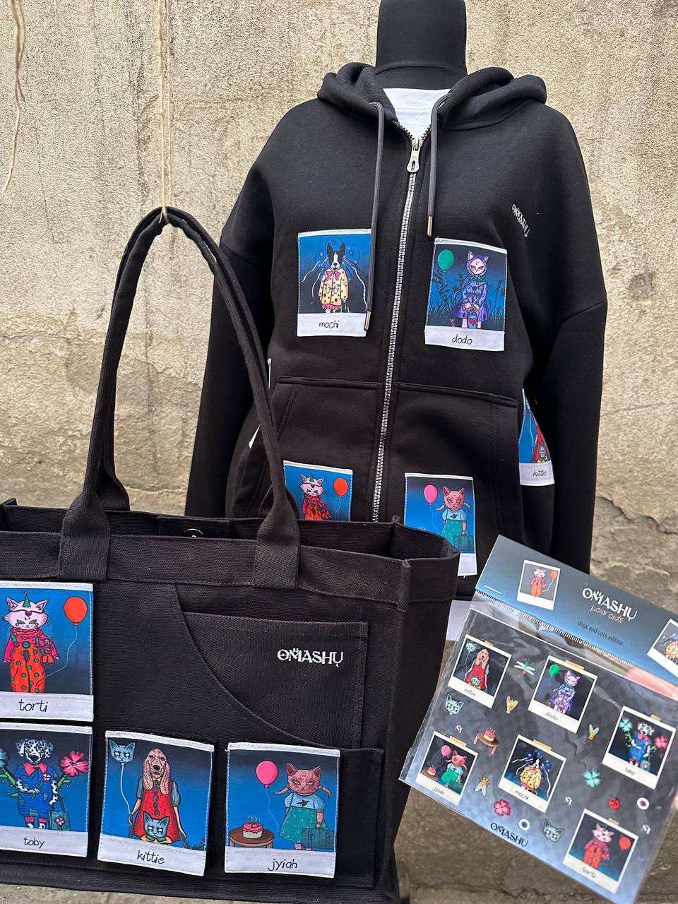 black merchandise from OMASHU featuring a hoodie and tote bag, both adorned with colorful Polaroid-style patches of animals. The hoodie displays multipale Polaroid patches of characters named "Mochi" and "Dodo," while the tote bag features additional characters like "Torti," "Toby," "Kittie," and "Jiyah." A display board featuring the same characters in a "dogs and cats edition" theme is also shown at the bottom right. The designs are vibrant and whimsical, with each animal wearing unique outfits and access