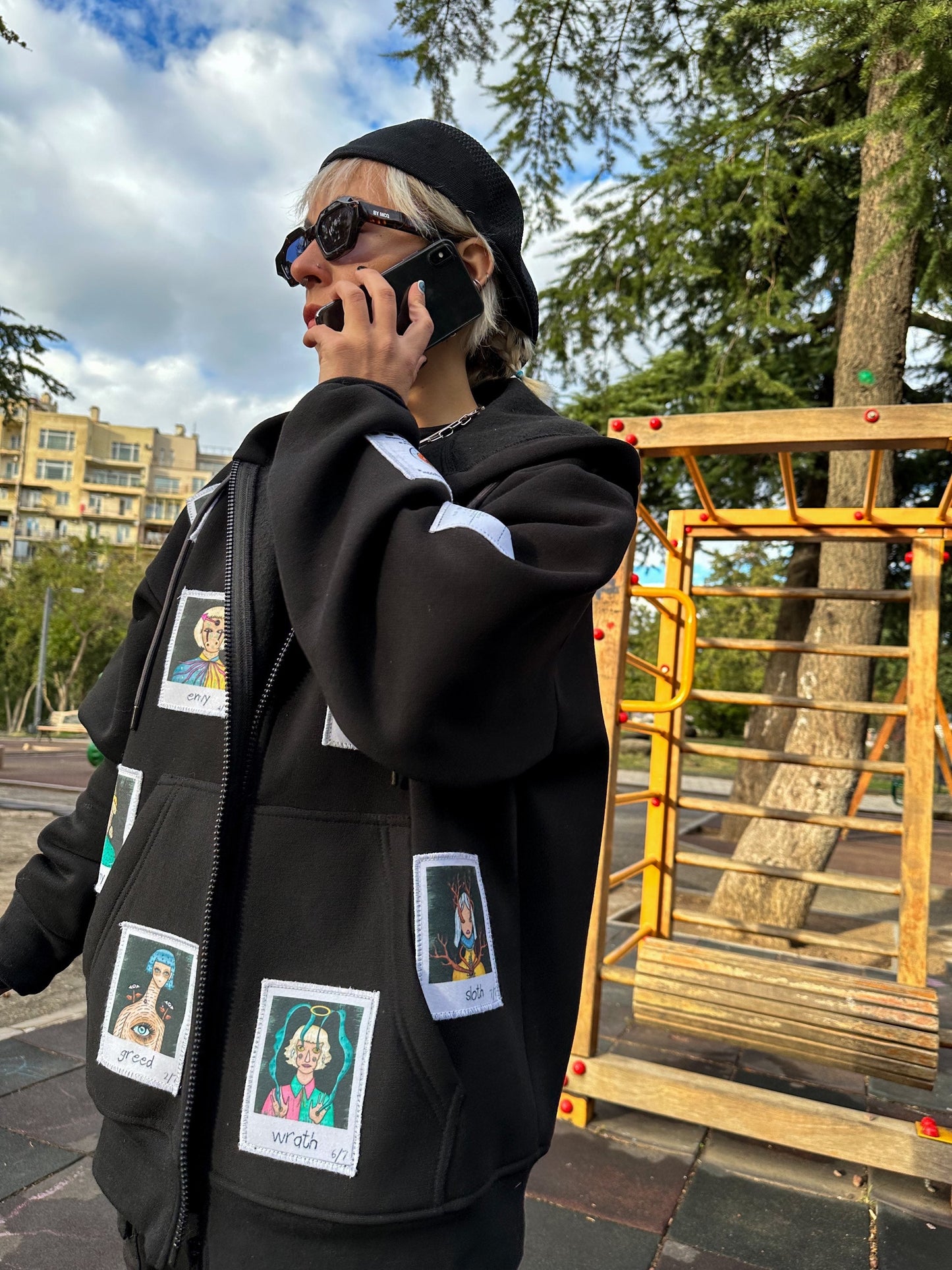 A person wearing a black hoodie with patches of different cartoon characters on it, a black baseball cap, and standing in a park with a jungle gym and a tree in the background.