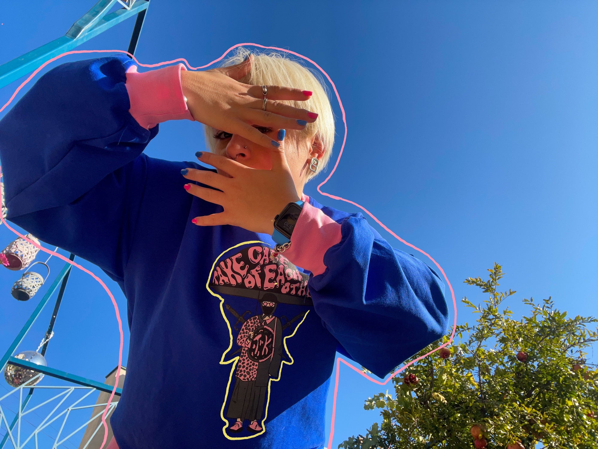 "A person wearing a blue sweatshirt with a graphic of a person holding a sign that reads ‘Take care of each other’ on it. The person is standing in front of a blue sky with a ferris wheel in the background. The person is covering their face with their hands, which have black nail polish on them. The person’s hair is blonde and styled in a bob. The photo is taken from a low angle, looking up at the person."1