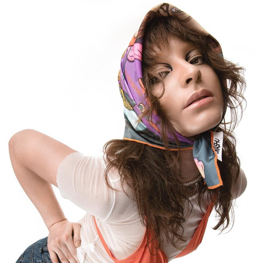 A photo of a person wearing a colorful headscarf with a pink and blue pattern, a white shirt, and an orange tank top, posing with their head tilted to the side and their hand on their hip, against a white background.