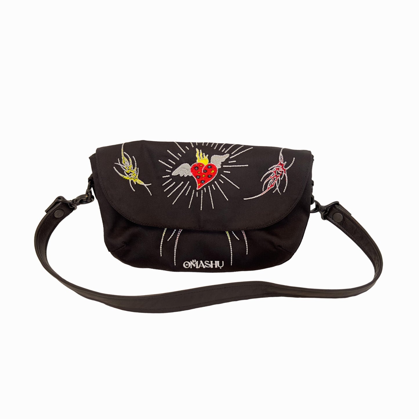A black messenger bag with a shoulder strap. The bag has a flap with a red heart, a yellow and red tattooesque figures The flap also has a white lines and the word ‘OMASHU’ written in white. The bag has a black shoulder strap with a black buckle.