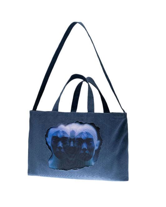 A blue tote bag with a cut-out window revealing a printed illustration, partially obscurred, against a light grey background.