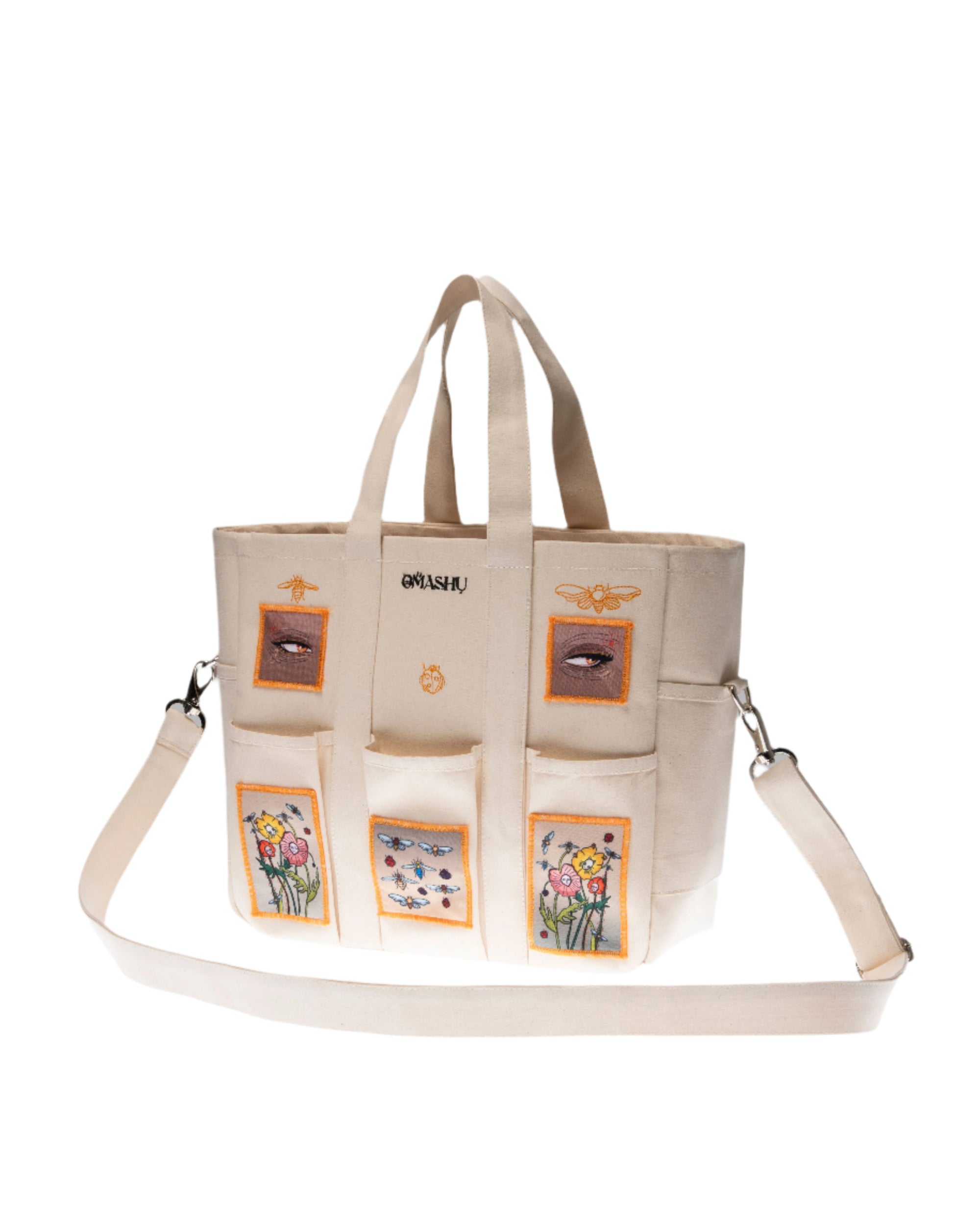 A beige tote bag with multiple pockets, each adorned with different embroidered designs including flowers and a crown, and the word ‘OWASHI’ stitched at the top, against a grey background.