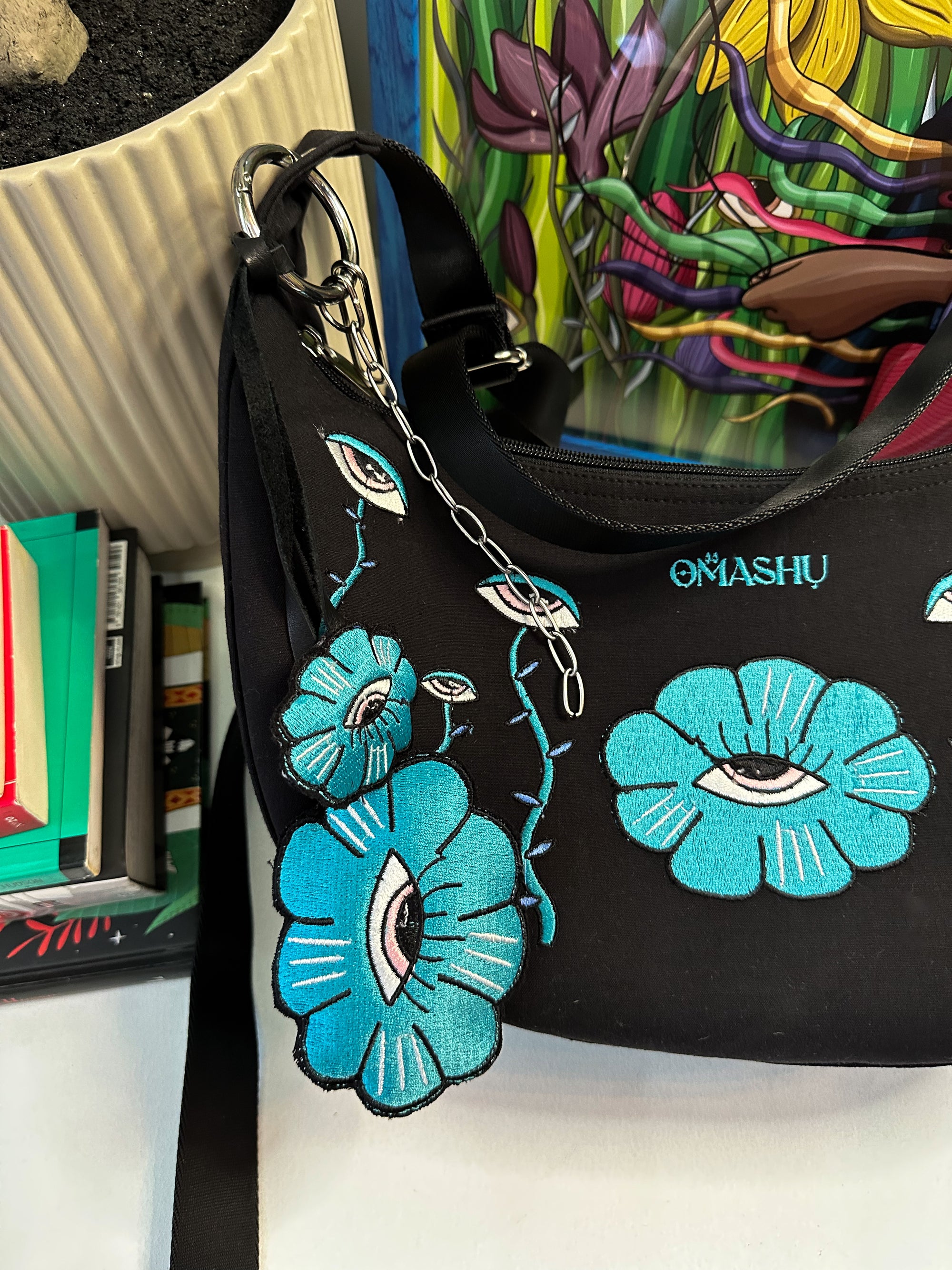 a bag and key chain both has embroidered illustration, it also has OMASHU branding on it. on the back ground there is a pot and books