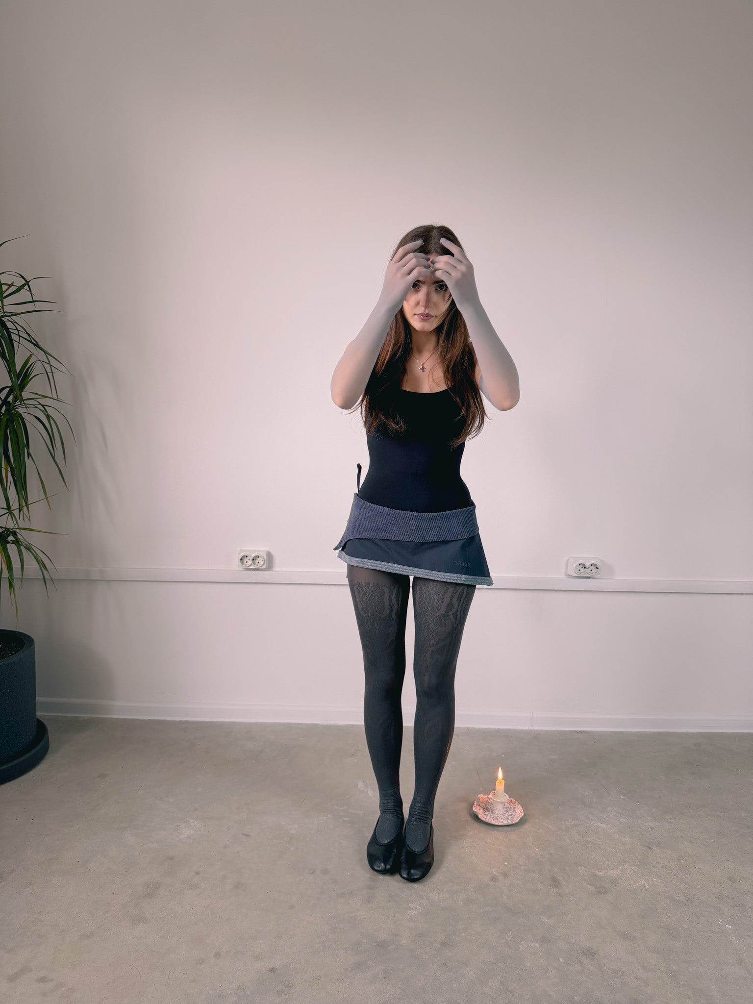 Person wearing a black top and denim skirt standing in a room with a white wall and a candle on the floor.