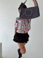 A person is wearing a colorful blouse with cartoon animal designs, including pandas and rabbits, along with a black ruffled skirt. The person is holding up a dark grey handbag with a large metal ring accent, obscuring part of their face. The playful outfit is paired with knee-high black socks, creating a fun and stylish combination. Visible tattoos on the legs add a unique detail to the overall look.