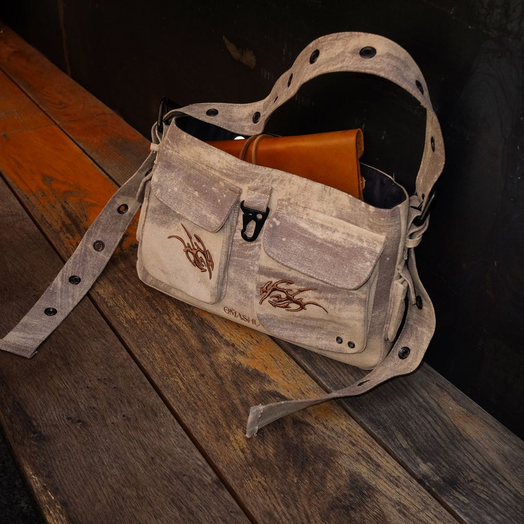A light gray, distressed canvas shoulder bag with a worn look, resting on a wooden surface. The bag features two front pockets with flap closures and brown tribal-style embroidery. The adjustable strap is accented with metal eyelets. The bag is partially open, revealing a brown leather item inside. The overall design has a rugged, vintage, and utilitarian style.