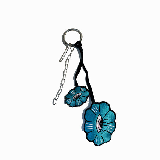 The keychain is made of a silver ring with a chain attached to it. The flowers are made of fabric and are attached to the chain with a black ribbon. The flowers are blue in color with a pink center and white details. The background is white.