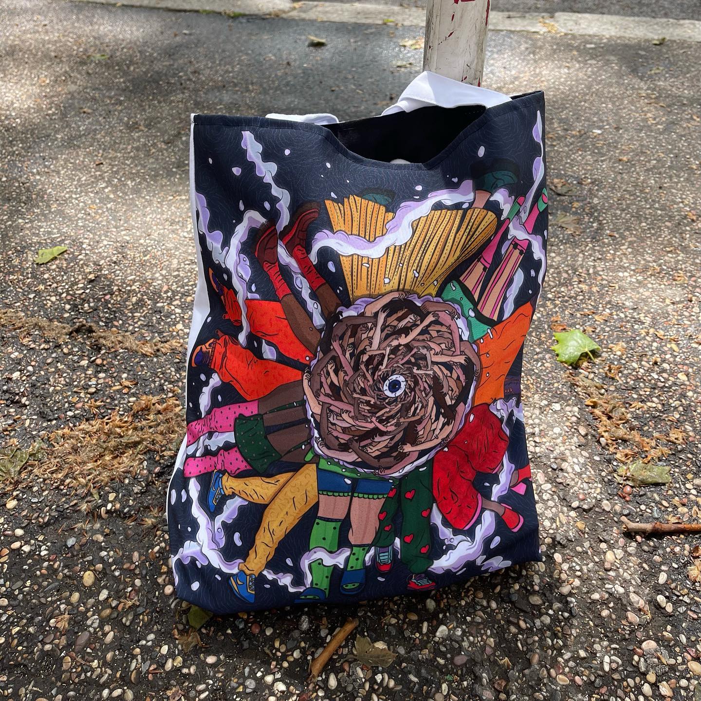 A black tote bag with a colorful abstract design on it. The design is a mix of different colors and shapes, including orange, yellow, green, and purple. The design appears to be a collage of different objects, including a bird, a flower, and a book. The tote bag is leaning against a tree trunk on a sidewalk with fallen leaves scattered around. The background is a street.