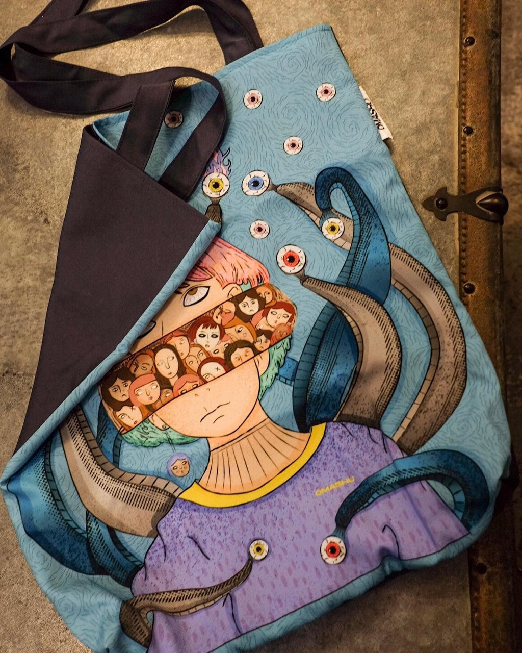 A colorful tote bag with an illustration of a person’s face with pink hair and a bandana covering their eyes. The bandana has illustrations of faces on it. The person’s face is surrounded by tentacles. The tote bag is made of a blue fabric with a swirling pattern on it and has a black strap and a triangular flap on the top.