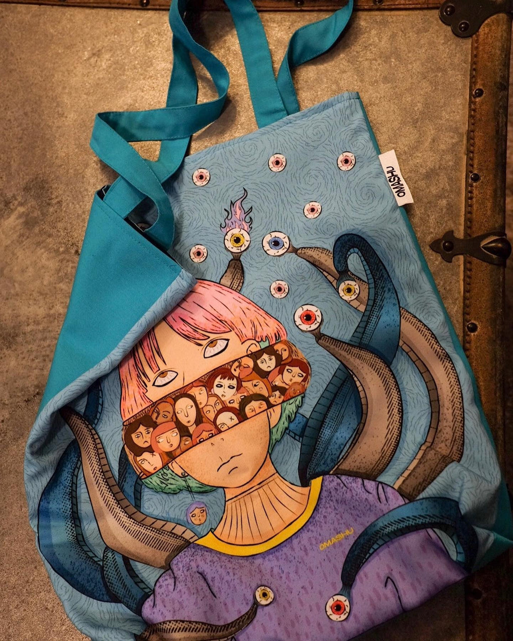 A blue tote bag with an illustration of a person with pink hair and a purple shirt, with tentacles and eyeballs surrounding them. The bag has a white tag on it that reads ‘OMASHU’. The background is a gray concrete wall with a metal hinge on the right side. The bag is hanging from a hook on the wall.