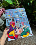Colorful illustration of building with gray color, blue windows, and pink roof with two characters in front of it, one red cat-like creature wearing yellow suit and one pink goat-like creature wearing green dress sitting on large orange cup with face on it.