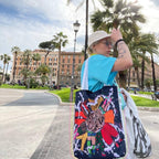 A person standing in a park with a large colorful tote bag. The person is wearing a blue t-shirt, white bucket hat, and carrying a white bag in addition to the tote bag. The tote bag is black with a colorful design of a tiger’s head and flowers. The background consists of a park with palm trees, a lamppost, and a building in the distance.