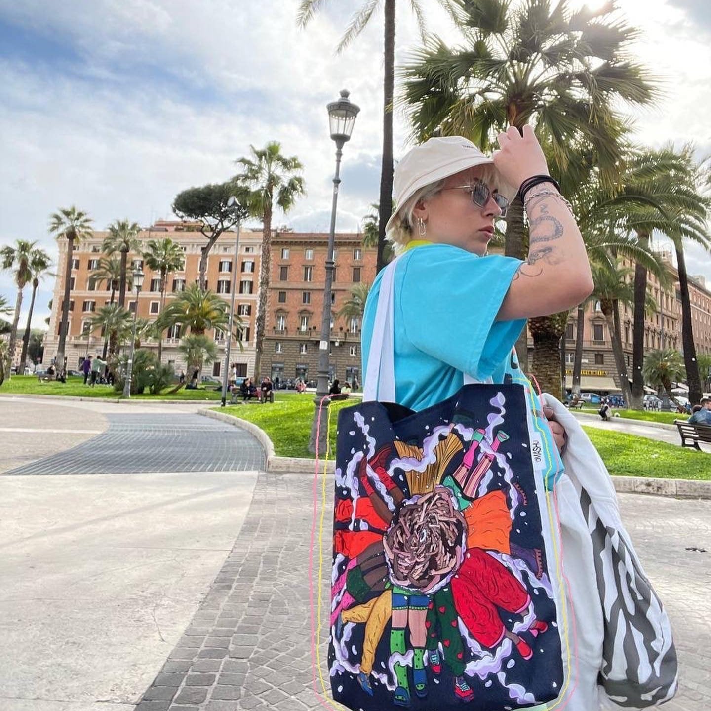 A person standing in a park with a large colorful tote bag. The person is wearing a blue t-shirt, white bucket hat, and carrying a white bag in addition to the tote bag. The tote bag is black with a colorful design of a tiger’s head and flowers. The background consists of a park with palm trees, a lamppost, and a building in the distance.