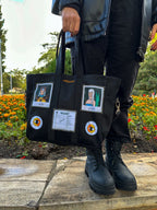 Black tote bag with various patches and pins arranged in a grid-like pattern. The patches include a cartoon polaroids and a receipt. The pins are round and have orange and yellow flowers on them. The person holding the bag is standing on a sidewalk with a flower bed in the background.