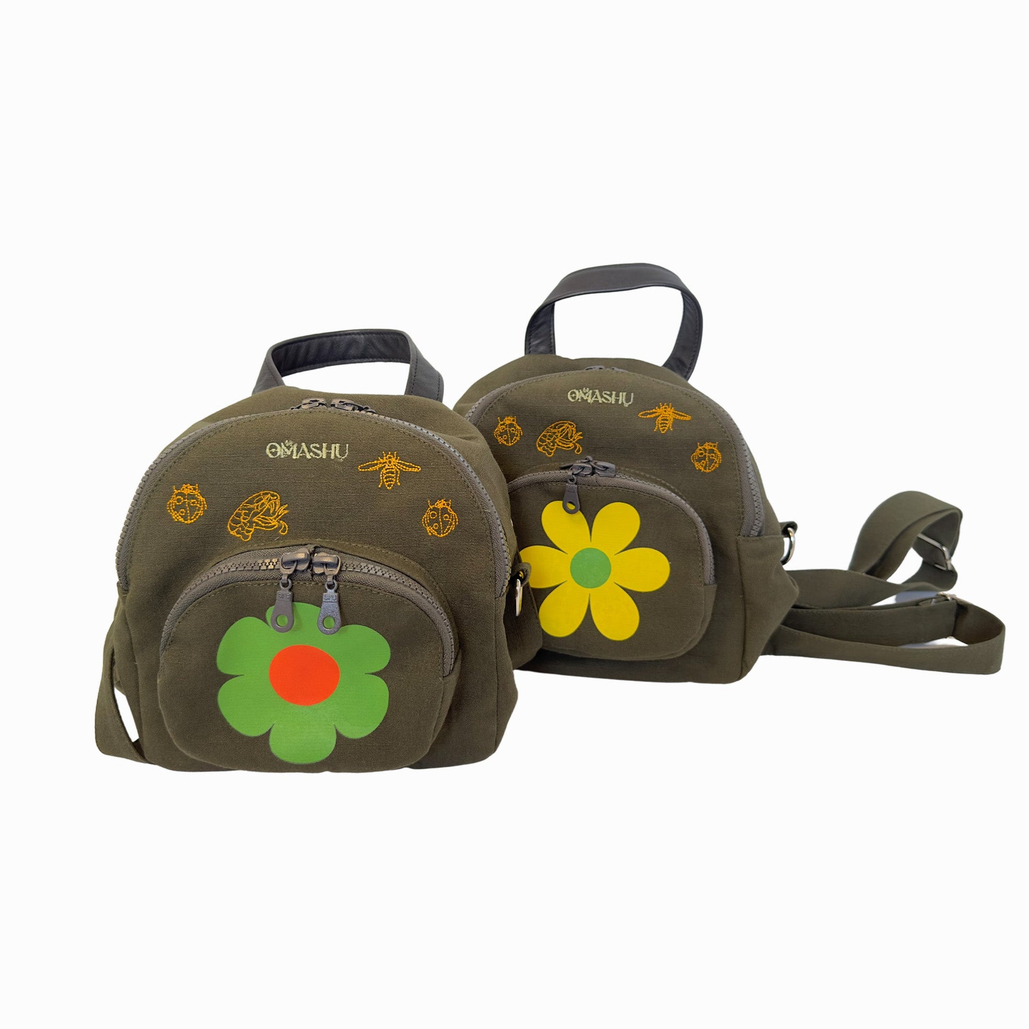 Two olive green backpacks with yellow and orange flower designs on them with black handle on top and black straps, zipper closure with green flower zipper pull, and the word ‘OMASHU’ written in yellow on top shown against white background.