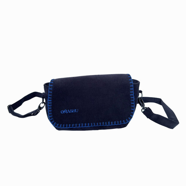 A photo of a black OMASHU crossbody bag with blue stitching, featuring a flap closure with a blue border, a black adjustable shoulder strap, and a small white logo on the flap that reads ‘OMASHU’.