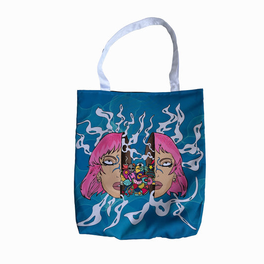 Omashu blue tote bag with a graphic design of pink-haired girl split in half facing each other with a in between them. The middle is filled with colorful candy and smoke is coming out of it. The girl has a blank expression on their faces. The background of the design is white with blue smoke swirling around it. The handles of the tote bag are white.