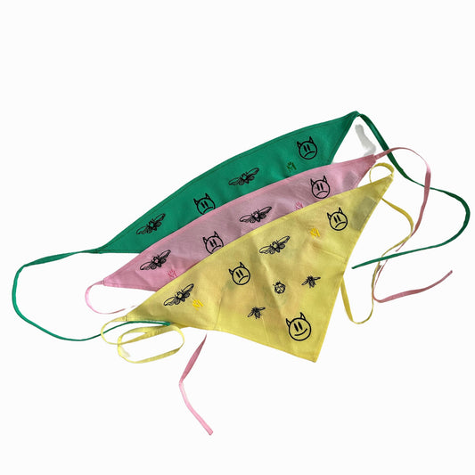 Three face scarves with a green and yellow panel. The scarves have black line drawings of insects and faces on them. The scarves are triangular in shape and have strings attached to them for tying around the head. The scarves are made of fabric. The masks are lying flat on a white background.